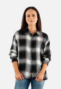 Camisa de franela a cuadros en blanco y negro con cuello redondeado, cierre de botones y sutiles adornos. Corte holgado con mangas arremangadas.