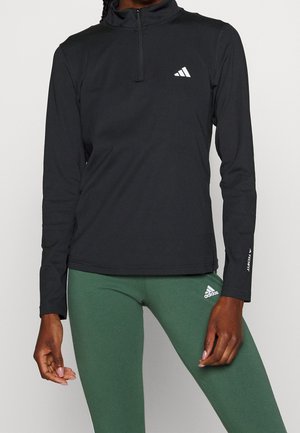 Personne portant un haut noir zippé à manches longues et un legging vert, les deux avec des logos blancs Adidas sur la poitrine et la cuisse.