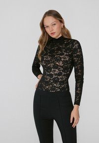 Top in pizzo nero a maniche lunghe con collo alto, caratterizzato da fantasie floreali e una silhouette aderente, abbinato a pantaloni neri a vita alta.