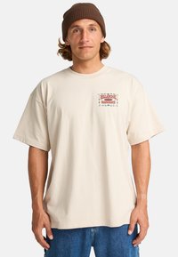 Beige cotton t-shirt with a front logo patch featuring red and blue text, short sleeves, and a relaxed fit. Worn with blue denim shorts.