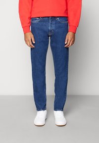 Vaqueros de mezclilla azul con corte recto, cinco bolsillos y detalles de costura sutiles, combinados con zapatillas deportivas blancas y una sudadera roja.