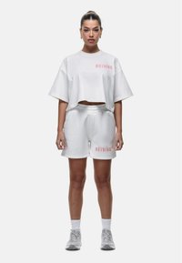 Cropped top in cotone bianco con testo stampato rosa, abbinato a pantaloni corti bianchi con testo coordinato. Il modello indossa sneaker e calzini bianchi.