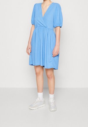 Femme portant une robe bleu clair mi-longue avec manches bouffantes, un décolleté plongeant en V, des chaussettes blanches courtes et des baskets blanches, debout sur un fond uni.