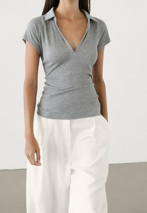 Kobieta ubrana w dopasowaną szarą krótką bluzkę z dekoltem w serek, włożoną do luźnych białych szerokich spodni, stojąca na jednolitym tle.