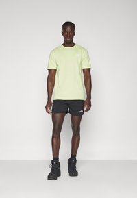Helles grünes T-Shirt mit kurzen Ärmeln und kleinem Logo, kombiniert mit schwarzen Shorts und schwarzen Wanderschuhen, steht vor einem schlichten Hintergrund.