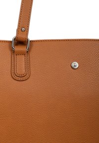 Sac fourre-tout en cuir brun avec une surface texturée, une bandoulière renforcée et un détail en argent sur le devant. Design simple et fonctionnel.