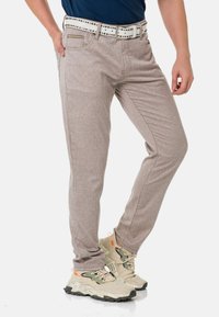 Hellbraune, strukturierte Slim-Fit-Hosen mit einem weißen Nieten-Gürtel. Kombiniert mit beigen Sneakers mit mehrfarbigen Akzenten.