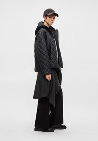 Melnā kvadrāta jakā ar kapuci, kas saskaņota ar garu melnu svārku un platu biksēm. Izskatu pilnveido melna cepure.