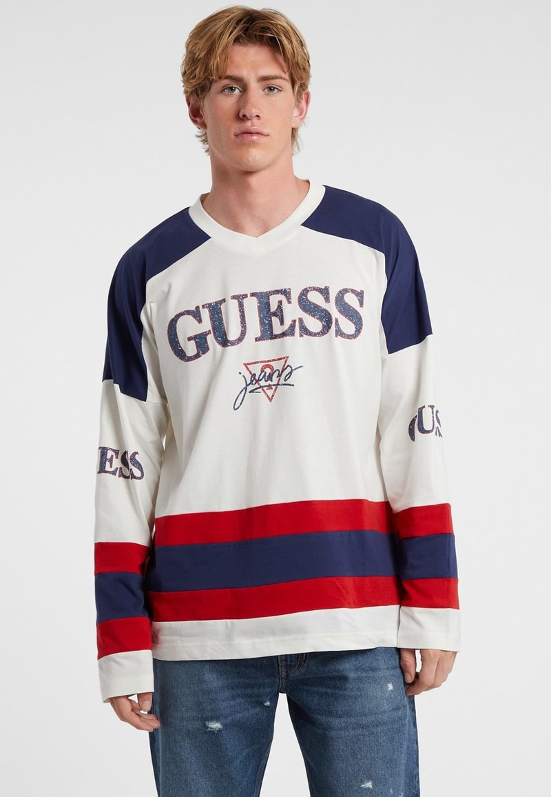 Langarm-T-Shirt in Creme mit horizontalen Streifen in Marineblau und Rot. Mit 'GUESS'-Logo auf der Vorderseite und den Ärmeln, aus weichem Baumwollstoff.