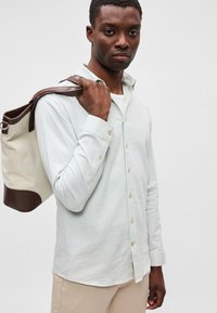 Φωτεινό πράσινο πουκαμίσo με μακριά μανίκια και χρυσά κουμπιά, φορεμένο με λευκή μπλούζα από κάτω. Μια μπεζ και καστανή τσάντα ώμου κρέμεται στον ώμο.