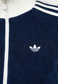 Casaco de cordura azul-marinho com fecho éclair e colarinho branco em malha. Apresenta detalhes em branco com três riscas e um pequeno logo branco da Adidas no peito.