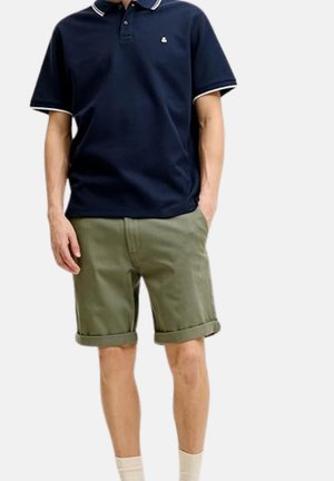 Hombre con camiseta polo azul marino de manga corta con ribete blanco y pantalones cortos verde oliva remangados, de pie con una mano en el bolsillo.