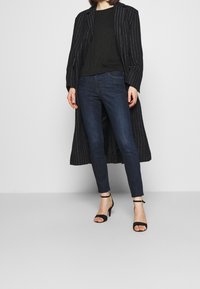 Kvinna i mörkblå slim fit jeans, svart topp, lång svart kappa med smala ränder och svarta högklackade sandaler med ankelremmar, stående på grått golv.