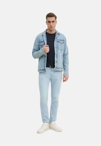 Giubbotto di jeans blu chiaro sopra una maglietta blu navy, abbinato a jeans blu chiaro e sneakers bianche. Il giubbotto ha tasche a pattina e una vestibilità casual.