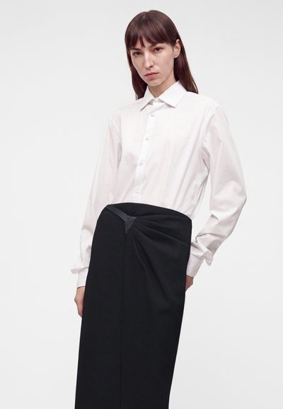 Femme portant une chemise blanche boutonnée rentrée dans une jupe noire taille haute avec un détail froncé à la taille, debout devant un fond uni.