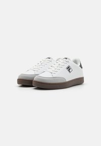 White sneakers with a smooth upper, grey suede toe cap, brown rubber sole, and black logo on the side. Features white laces and padded collar.