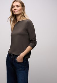 Brauner Langarmshirt mit lockerem Schnitt, rundem Ausschnitt und strukturierter Stoffqualität, kombiniert mit dunkelblauen Jeans mit aufgesetzten Fronttaschen.