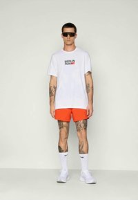Muž s bielym tričkom "Berlin Run", jasne oranžovými šortkami, bielymi ponožkami a topánkami Nike, tmavými slnečnými okuliarmi, stojaci s viditeľnými tetovaniami na rukách a nohách.
