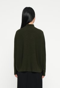 Donna con i lunghi capelli neri che indossa un maglione verde scuro e una gonna nera a pieghe, in piedi di spalle contro uno sfondo semplice.