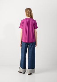 Fuksia lyhythihainen paita, jossa on kokoontunut selkä, yhdistettynä leveälahkeisiin sinisiin farkkuihin, joissa on harmaa purjehtiva helma. Yhdistetty litteisiin sandaaleihin.