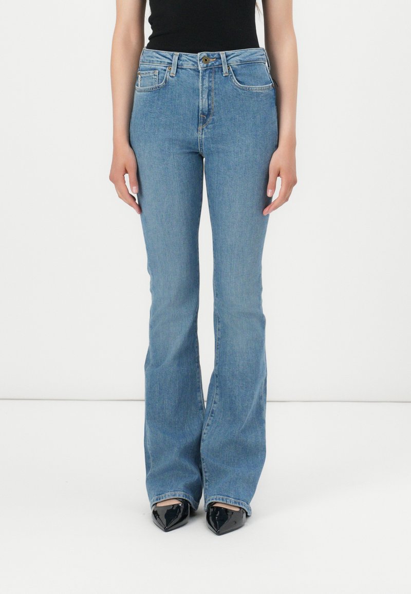 Calças de ganga claras, com corte à boca de sino e cintura alta, apresentando bolsos frontais e fecho de botão. Textura de ganga suave, combinadas com sapatos de salto pretos de bico.