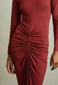 Abito a maniche lunghe di color ruggine realizzato in tessuto elasticizzato, con arricciature verticali lungo il torso che creano texture e forma.