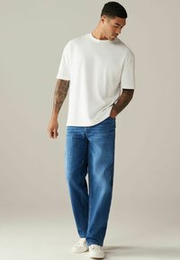 Tricou alb cu mânecă scurtă, croială lejeră, asociat cu blugi albastri drepți și teniși albi. Design simplu, fără modele sau logo-uri vizibile.