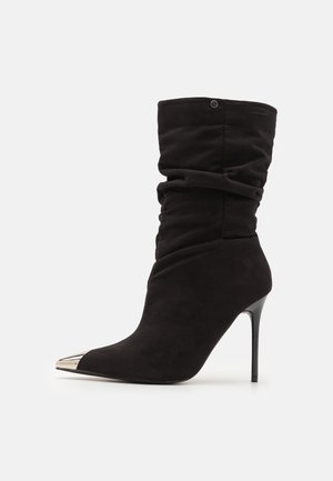 Bottine noire décontractée en daim avec un bout pointu et une coiffe métallique. Caractéristiques d'un talon haut stilettos et d'une texture froncée autour de la tige.
