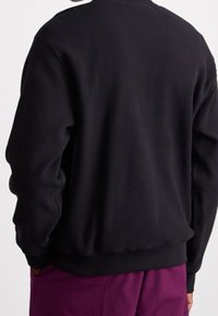 Persona che indossa una maglietta nera a maniche lunghe con textura e pantaloni viola, mani parzialmente nelle tasche, con la schiena rivolta verso la macchina fotografica.
