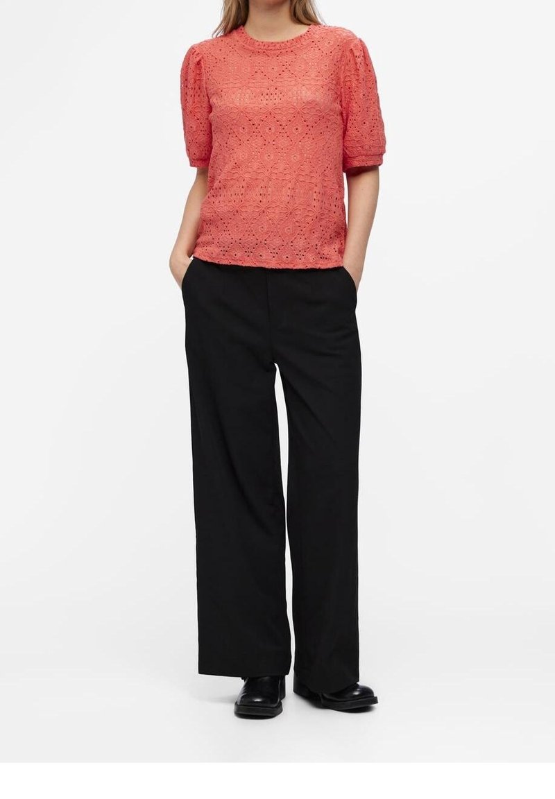 Femme portant un top court en dentelle corail, un pantalon noir à jambes larges et des chaussures noires, debout les mains dans les poches.
