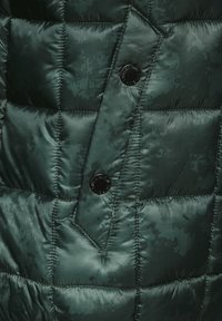 Groene gewatteerde jasstof met een glanzende afwerking, voorzien van een kenmerkend camouflagepatroon. Opvallende zwarte knoopaccent nabij de plooi.