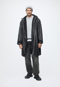 Cămașă lungă, impermeabilă, de culoare neagră, cu căptușeală din fleece moale, hanorac gri dedesubt, blugi de culoare gri închis și pantofi sport negri. Căciulă gri.