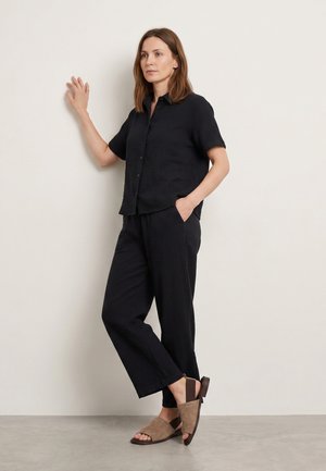 Vrouw staat tegen een muur, draagt een zwart overhemd met korte mouwen en knopen, zwarte broek en beige sandalen met open tenen.
