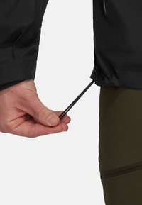 Giacca nera impermeabile con polsini regolabili, dotata di un cordoncino di serraggio e una superficie liscia. Mano che afferra il cordoncino vicino a un pantalone verde oliva.