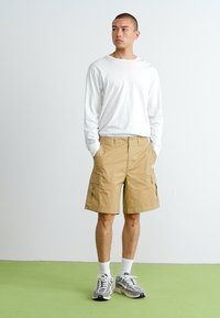 Calções cargo bege com múltiplos bolsos laterais, combinados com uma camisa branca de mangas compridas e sapatilhas cinzentas num piso verde. Design simples.