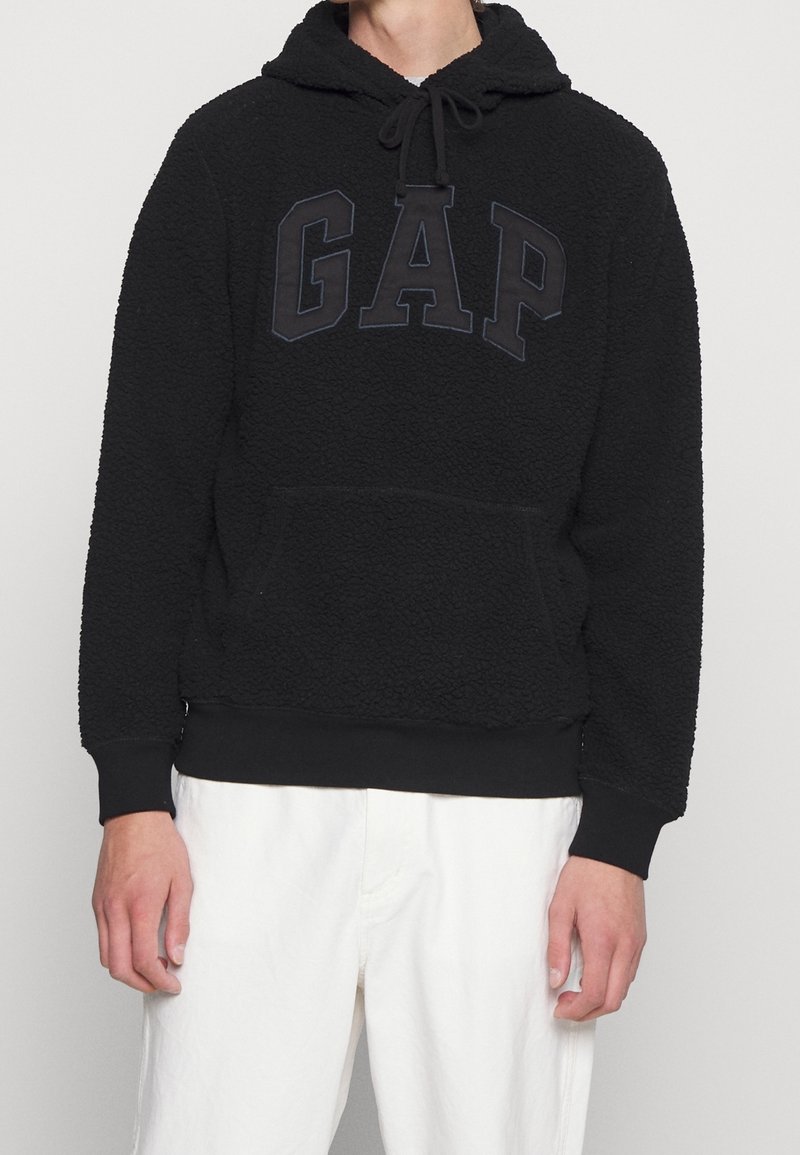 Personne portant un sweat à capuche GAP noir texturé avec poche avant et cordons noirs, associé à un pantalon blanc.