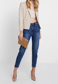 Blazer beige, top court en dentelle, jeans ajustés bleu marine, escarpins nude et une pochette dorée froissée, mettant en valeur des textures et des formes variées.