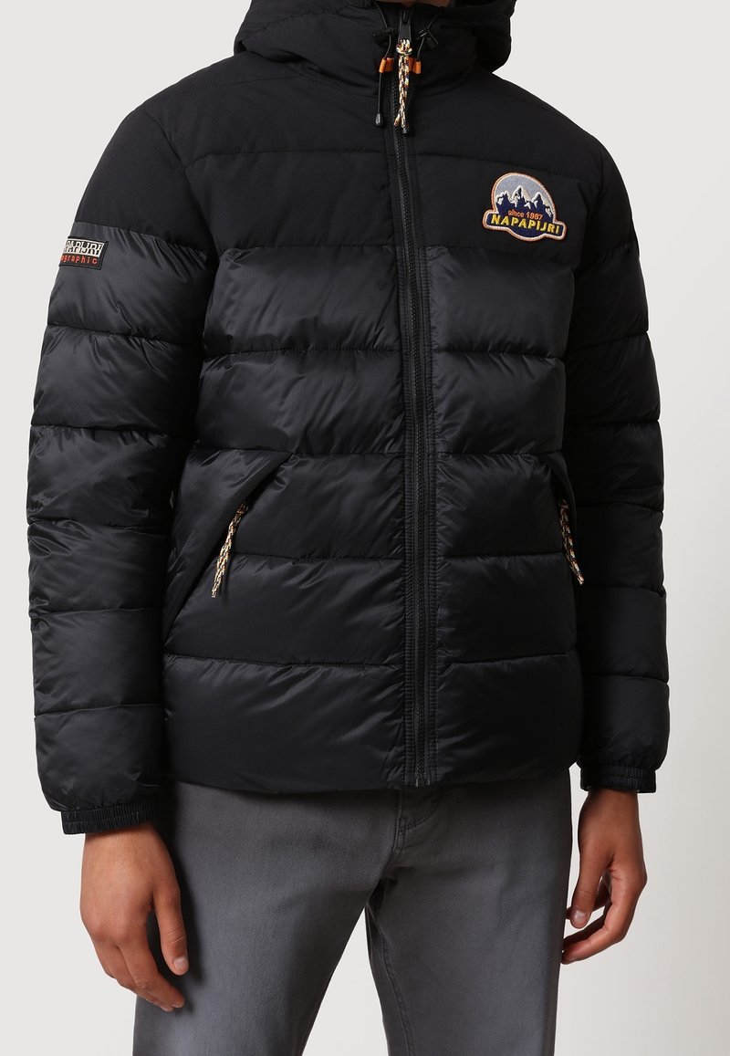 Veste matelassée noire à capuche avec fermeture éclair, écussons de marque sur la poitrine et la manche, et poches latérales zippées, portée par une personne avec un pantalon gris.