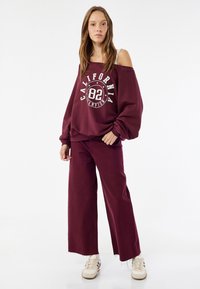 Bordeaux-huoneen oversize-sweatshirt, jossa on valkoinen graafinen printti ja paljaat hartiat, yhdistettynä samanvärisiin leveisiin burgundihousuihin ja kermaisiin tennareihin.