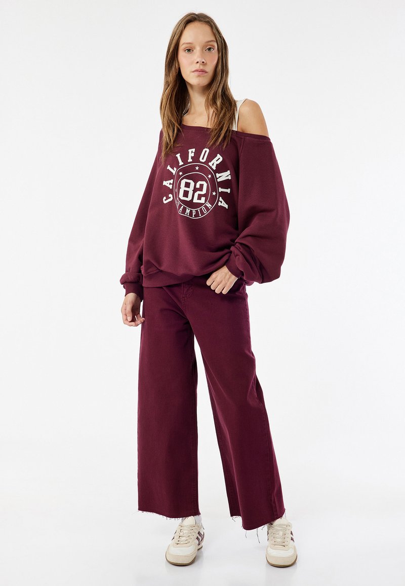 Bordeaux-huoneen oversize-sweatshirt, jossa on valkoinen graafinen printti ja paljaat hartiat, yhdistettynä samanvärisiin leveisiin burgundihousuihin ja kermaisiin tennareihin.