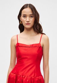 Κόκκινο satino φόρεμα με λεπτές τιράντες, πλισέ φούστα και τετράγωνη λαιμοκοπή. Λείος υφασμάτινος ιστός με διακριτή γυαλάδα και χωρίς ορατά μοτίβα.