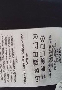 Eticheta cu instrucțiuni de îngrijire imprimată în negru pe un material alb. Include simboluri pentru spălare, uscare și tehnici de călcare.