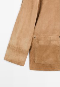 Giacca in suede color marrone chiaro, con maniche ampie, dettagli cuciti e una grande tasca a toppa con accenti di hardware visibili.