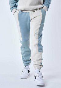 Los pantalones de chándal en azul claro y crema presentan un diseño de bloques de color, puños elásticos y detalles bordados. Combinados con zapatillas blancas.