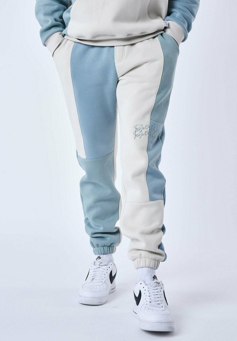 Los pantalones de chándal en azul claro y crema presentan un diseño de bloques de color, puños elásticos y detalles bordados. Combinados con zapatillas blancas.