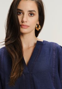 Granatowa lniana bluzka z dekoltem w serek, z koronkowymi detalami z przodu. Modelka nosi złote kolczyki w kształcie kół, a włosy ma rozpuszczone.