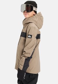 Mladý človek oblečený v béžovo-čiernej lyžiarskej bunde, čiernej čiapke, veľkých reflexných lyžiarskych okuliaroch a čiernych rukaviciach, stojaci bokom na bielom pozadí.