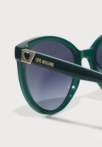 Roheline plastist päikeseprillid halli gradient-läätsedega. Omaduseks on kuldse tooni detailid südame kujutisega ja "LOVE MOSCHINO" kiri käepideme peal.