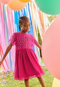 Roze jurk met korte, geborduurde mouwen en bloemenmotieven. Uitlopende rok, lichtgewicht stof. Achtergrond met kleurrijke ballonnen en slingers.