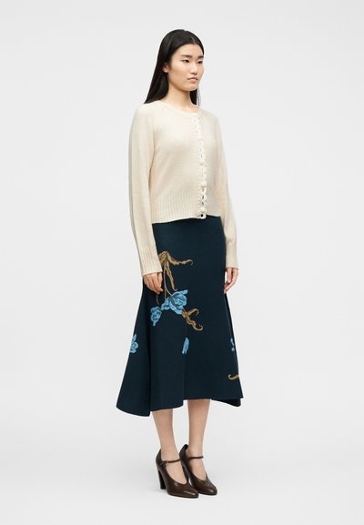 Γυναίκα που φοράει κρεμ ζακέτα με κουμπιά, σκούρα μπλε μίντι φούστα με μπλε και καφέ κεντήματα λουλουδιών, και καφέ ψηλοτάκουνα παπούτσια τύπου Mary Jane.